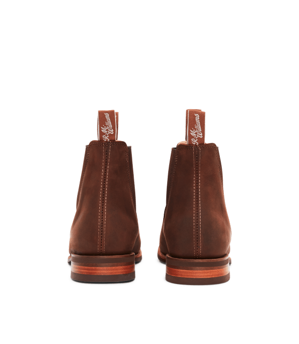 A pair of brown leather Chelsea boots viewed from the back, showing pull tabs with white script and visible stitching down the centre.