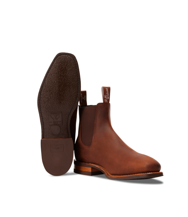 A pair of brown leather Chelsea boots, one boot standing upright and the other lying on its side to display the textured sole and heel on a plain white background.