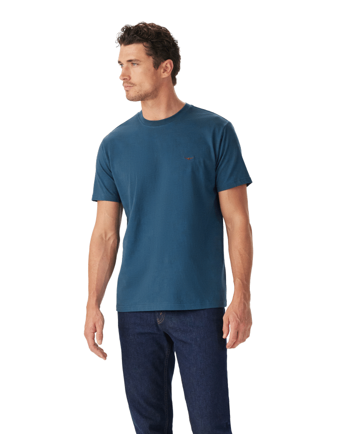 A man stands facing left, wearing a blue short-sleeve t-shirt and dark blue jeans, against a plain white background.