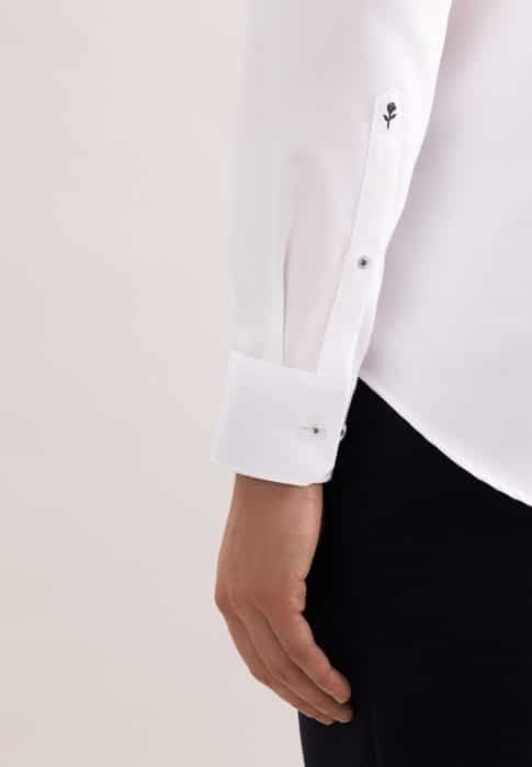 A close-up of a person’s arm wearing a white long-sleeved shirt with buttoned cuffs and black trousers. The shirt sleeve features a small embroidered black symbol near the wrist.