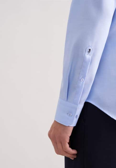 A person wearing a light blue long-sleeved dress shirt with buttoned cuffs and black trousers. The shirt has a small embroidered detail near the sleeve vent. The background is plain and light-coloured.