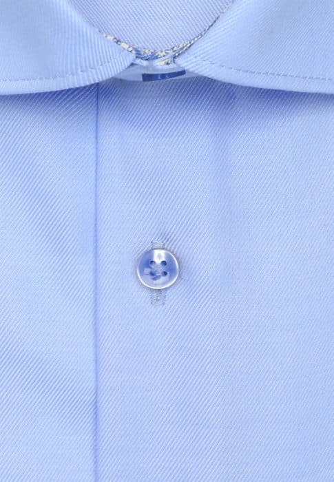 Close-up of a light blue dress shirt, showing a translucent button sewn onto the fabric with a diagonal weave pattern. The top of the shirt’s collar and neckline are also partially visible.