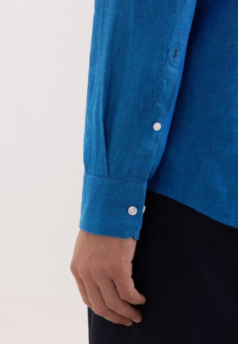 A close-up of a person’s arm wearing a bright blue long-sleeve shirt with white buttons on the cuff. The hand is relaxed by their side, and the background is plain and light-coloured.