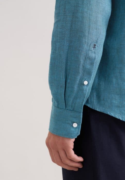 A close-up of a person's arm wearing a long-sleeved, teal-coloured button-up shirt with white buttons and dark trousers, shown from the side against a plain background.