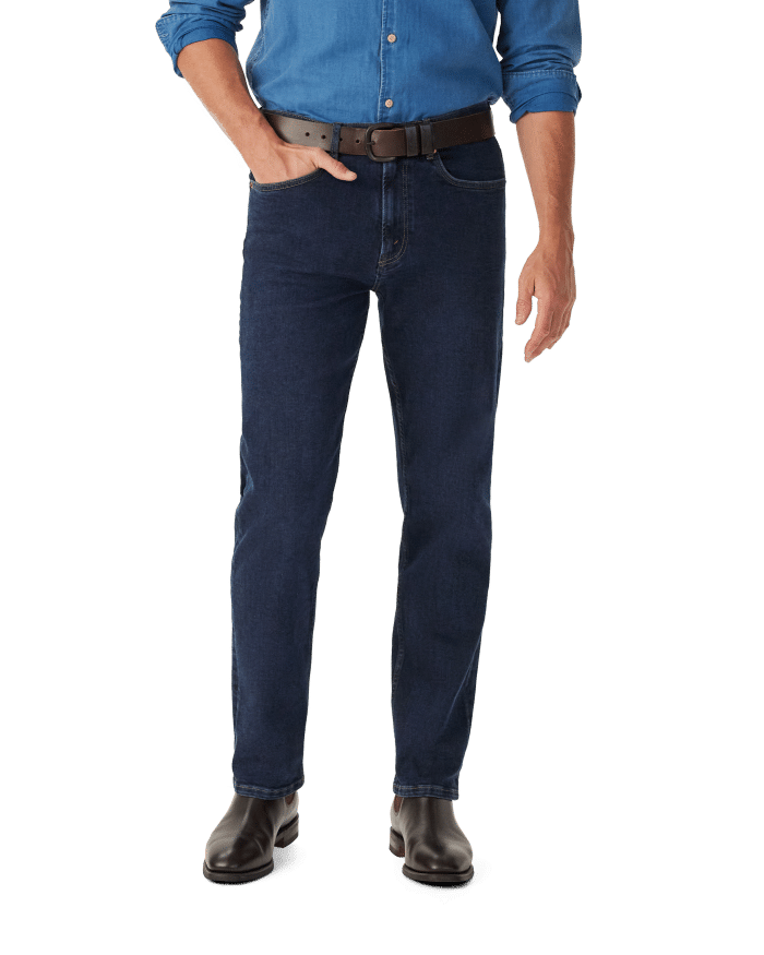 A person wearing a blue denim shirt, dark blue jeans, a brown belt, and brown shoes stands with one hand in their pocket against a plain white background.