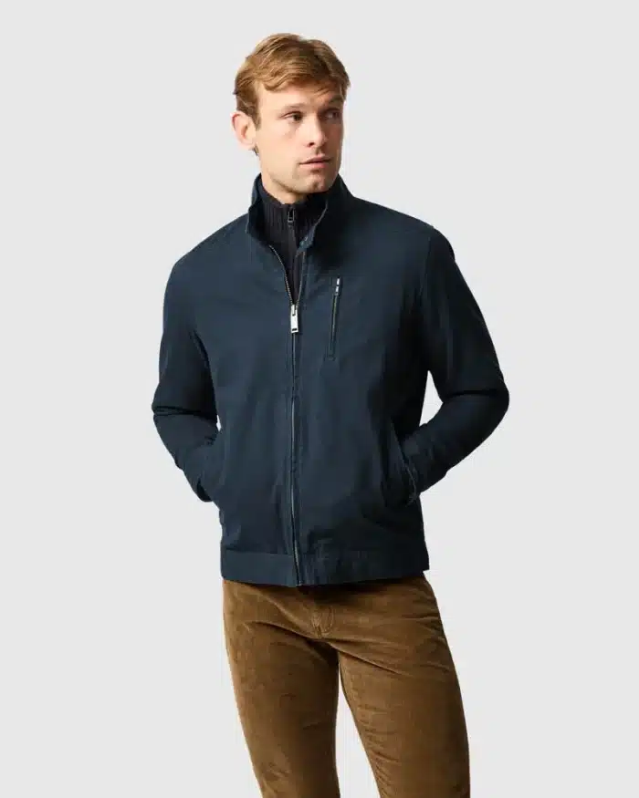 A man wearing a navy blue zip-up jacket over a dark top and brown corduroy trousers stands with his hands in his jacket pockets, looking to the side against a plain light grey background.