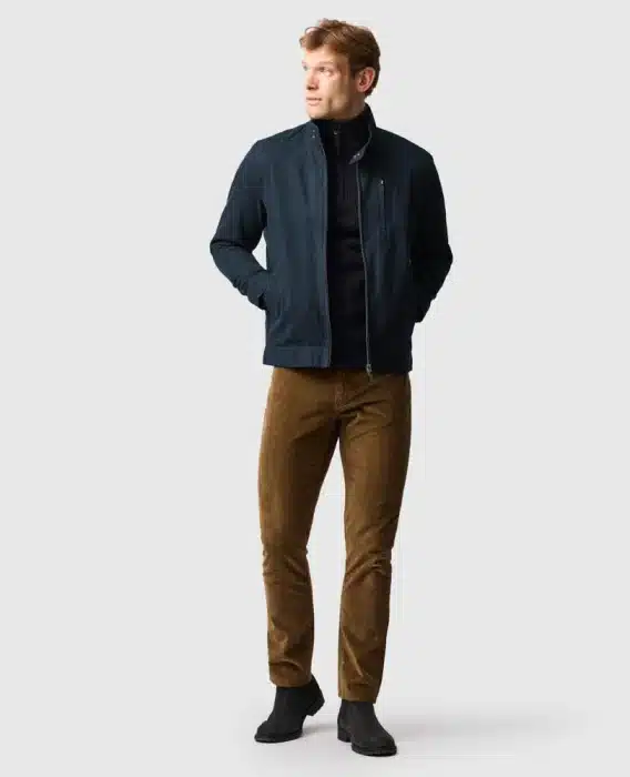 A man stands with hands in his jacket pockets, wearing a navy jacket, dark jumper, brown corduroy trousers, and black shoes against a plain light grey background.