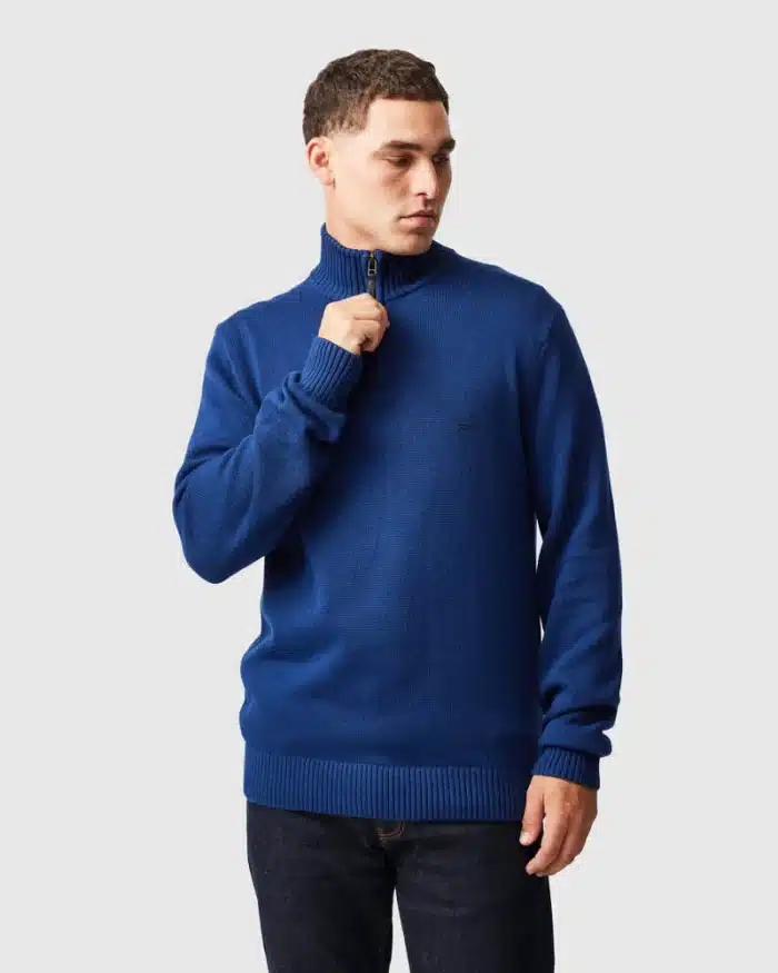 A man wearing a royal blue zip-up mock neck jumper and dark jeans stands against a plain light background, looking to his left whilst holding the jumper’s zip near his chin.