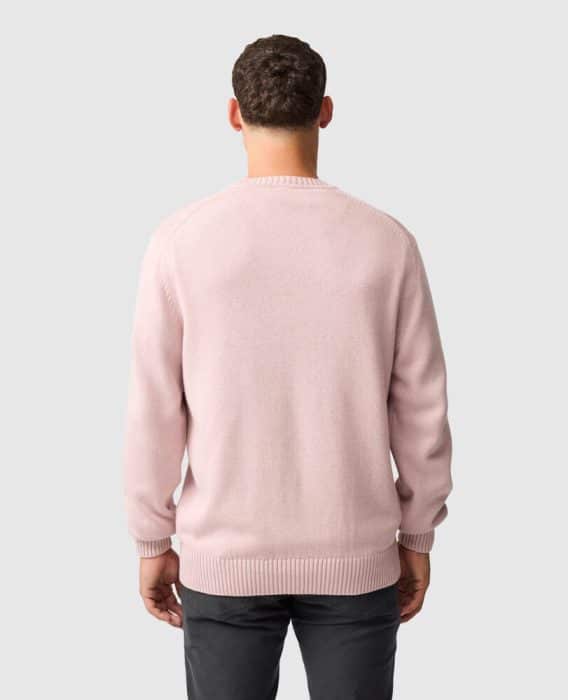 A man with short brown hair wearing a light pink jumper and dark trousers stands facing away, shown from the back against a plain light grey background.