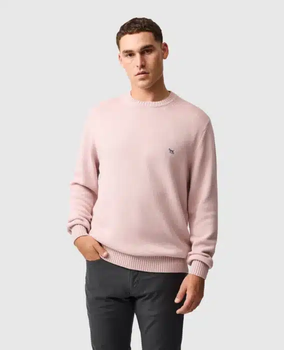A man stands against a plain light grey background, wearing a pale pink jumper with an embroidered logo on the chest and dark grey trousers. He has short hair and a neutral expression, with one hand in his pocket.