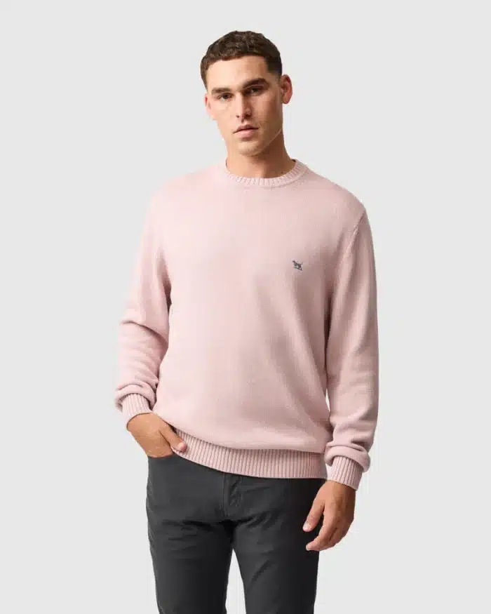 A man stands against a plain light grey background, wearing a pale pink jumper with an embroidered logo on the chest and dark grey trousers. He has short hair and a neutral expression, with one hand in his pocket.