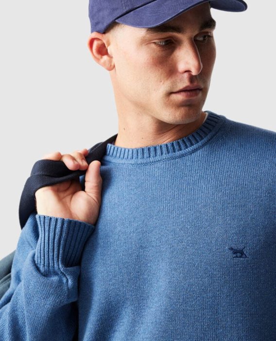 A man wearing a blue baseball cap and a blue jumper with a small embroidered logo looks to the side whilst holding a jacket over his shoulder.