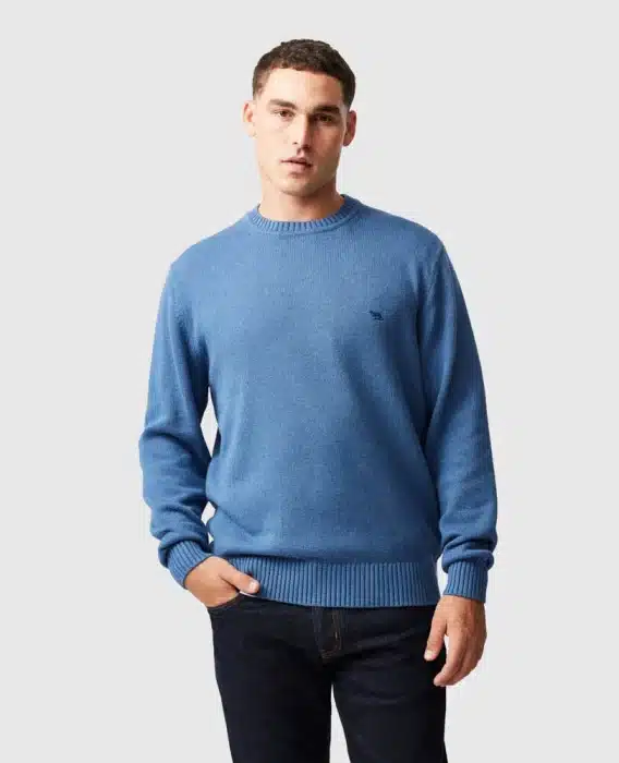 A man wearing a light blue crew neck jumper and dark jeans stands against a plain light grey background with one hand in his pocket, looking at the camera.