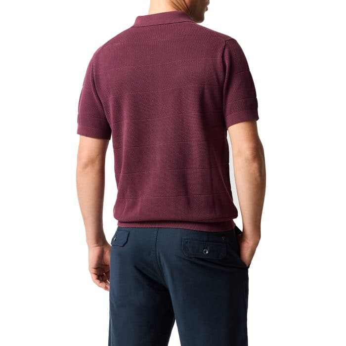 A man stands facing away, wearing a short-sleeve maroon polo shirt and navy blue trousers against a white background.