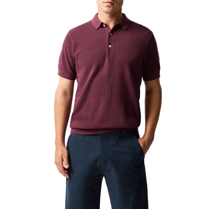 A man wearing a short-sleeve, maroon polo shirt and dark navy trousers stands with one hand in his pocket against a white background.
