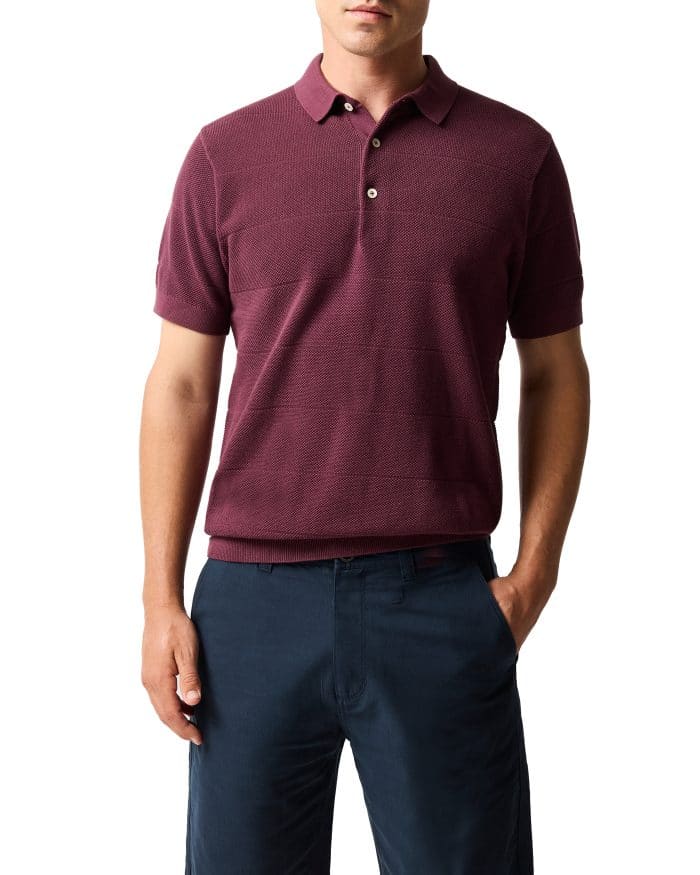 A man wearing a short-sleeve, maroon polo shirt and dark navy trousers stands with one hand in his pocket against a white background.
