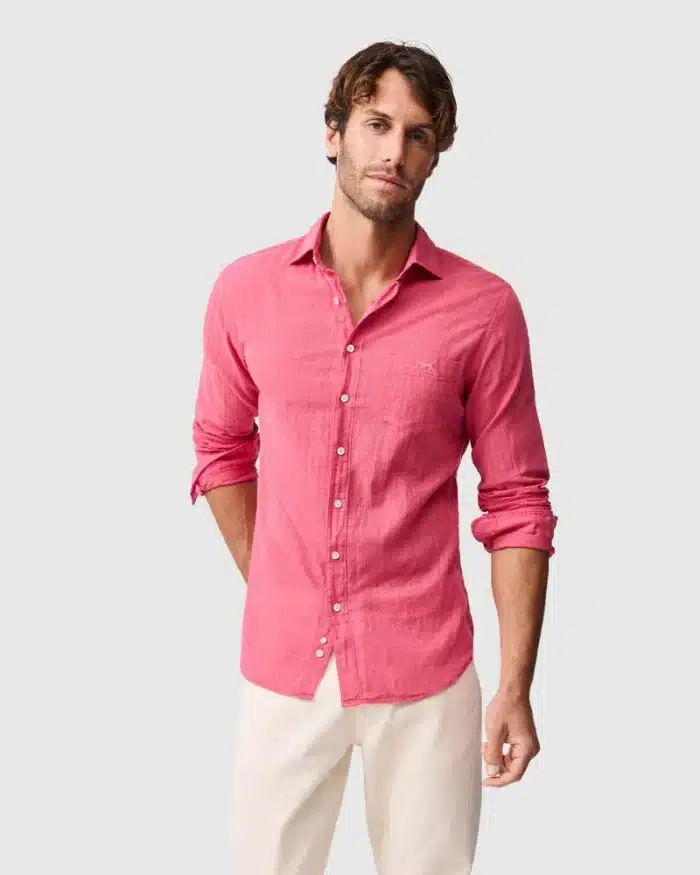A man wearing a long-sleeved, pink button-up shirt and white trousers stands against a plain light grey background with his sleeves rolled up and one hand in his pocket.