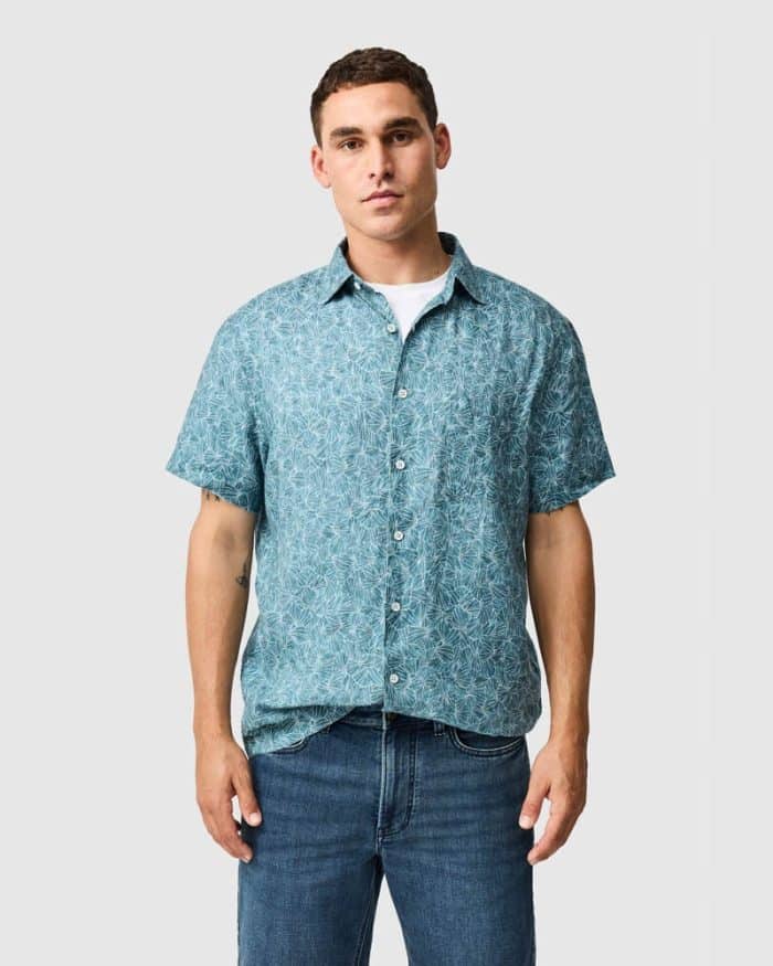 A man stands facing forward, wearing a light blue short-sleeve button-up shirt with a white geometric pattern and blue jeans, over a white vest, against a plain light grey background.