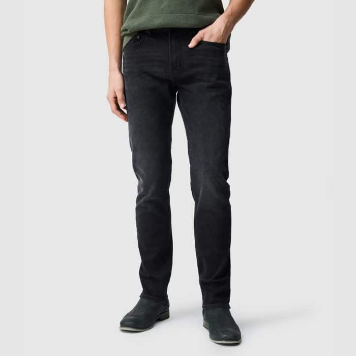 A person wearing a green shirt, black jeans, and dark shoes stands against a light grey background, with their arms relaxed at their sides.