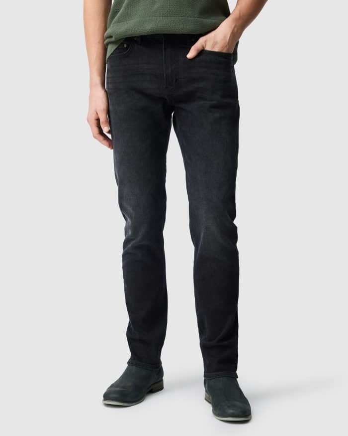 A person wearing a green shirt, black jeans, and dark shoes stands against a light grey background, with their arms relaxed at their sides.