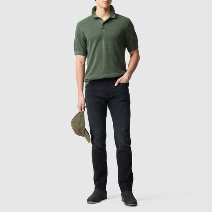 A man wearing a green polo shirt, black jeans, and dark shoes stands holding a green cap by his side against a plain light grey background.