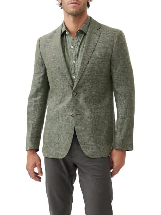 A man wears a green textured blazer over a green button-up shirt and dark trousers. The outfit is stylish and casual, and the background is plain white.