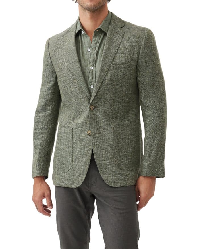 A man wears a green textured blazer over a green button-up shirt and dark trousers. The outfit is stylish and casual, and the background is plain white.