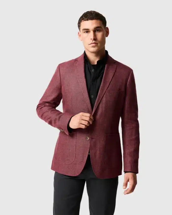 Man wearing a maroon blazer over a black button-up shirt and dark trousers stands against a plain light grey background, looking at the camera with a neutral expression.