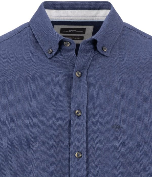Close-up of a blue button-up shirt with a subtle herringbone pattern, button-down collar, and embroidered logo on the left chest. Four buttons are visible on the placket.