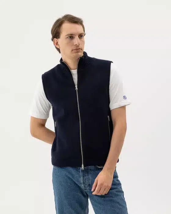 A person wearing a navy zip-up waistcoat over a white T-shirt and blue jeans stands against a plain white background, looking slightly to the side.