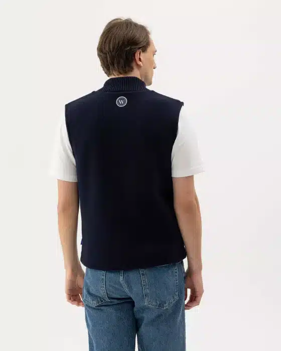 A person with short brown hair, wearing a white T-shirt, a dark sleeveless jumper with a small circular logo on the back, and blue jeans, stands facing away against a plain white background.