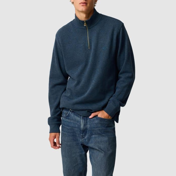 A person wearing a dark blue quarter-zip jumper and blue jeans stands with one hand in their pocket against a plain light grey background.