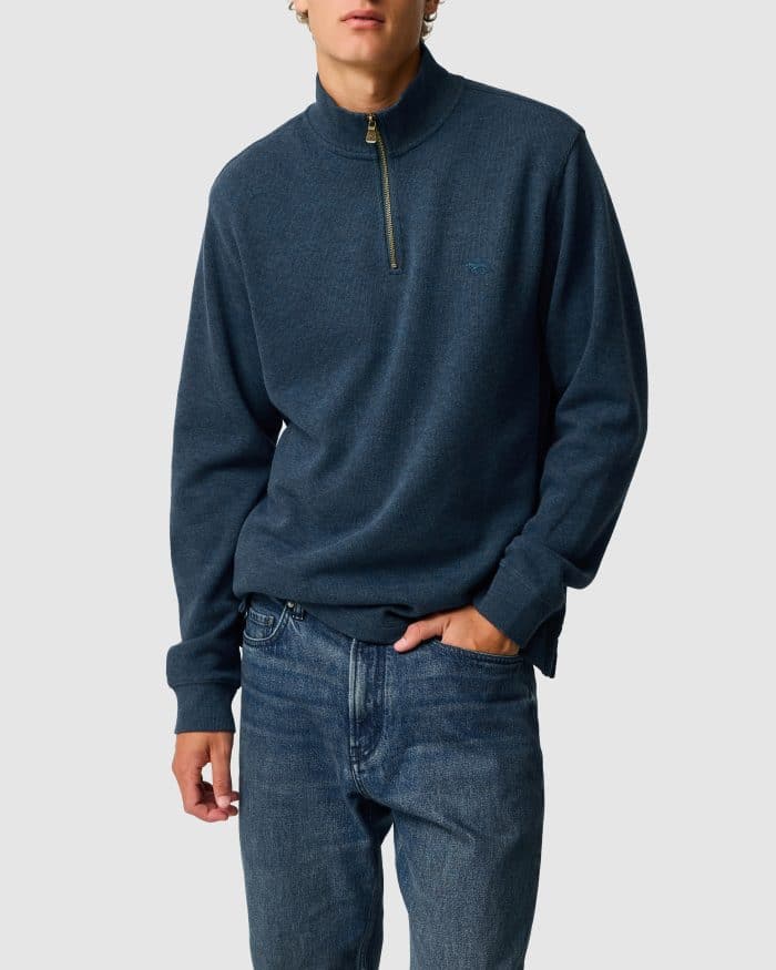 A person wearing a dark blue quarter-zip jumper and blue jeans stands with one hand in their pocket against a plain light grey background.