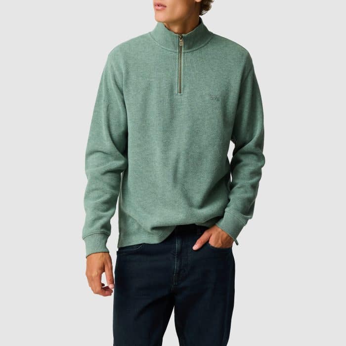 A person wearing a light green quarter-zip jumper and dark jeans stands against a plain light grey background. Their head is turned slightly to the side and one hand is in their pocket.