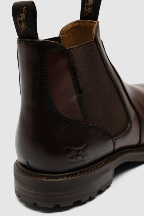 Close-up of the back and side of a dark brown leather Chelsea boot with elasticated panels and pull tabs featuring a decorative emblem. The boot has a sturdy sole and subtle stitching details.