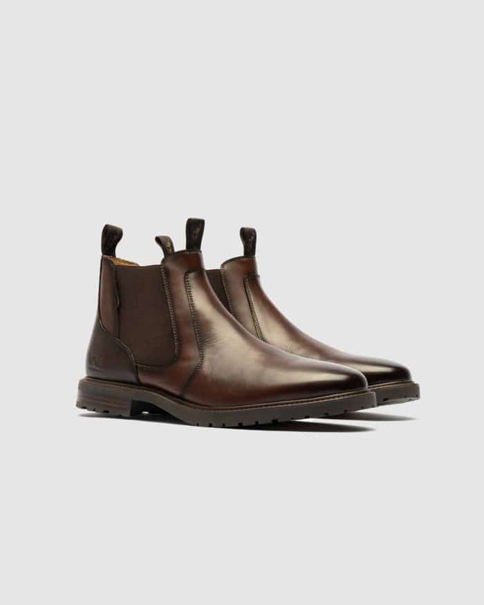 Pair of brown leather Chelsea boots with elastic side panels and pull tabs, displayed on a light grey background. The boots have a sleek, polished finish and slightly pointed toes.