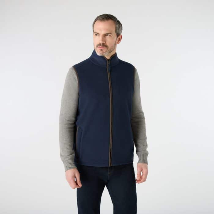 A man wearing a dark blue zip-up fleece gilet over a grey long-sleeved shirt and dark jeans.