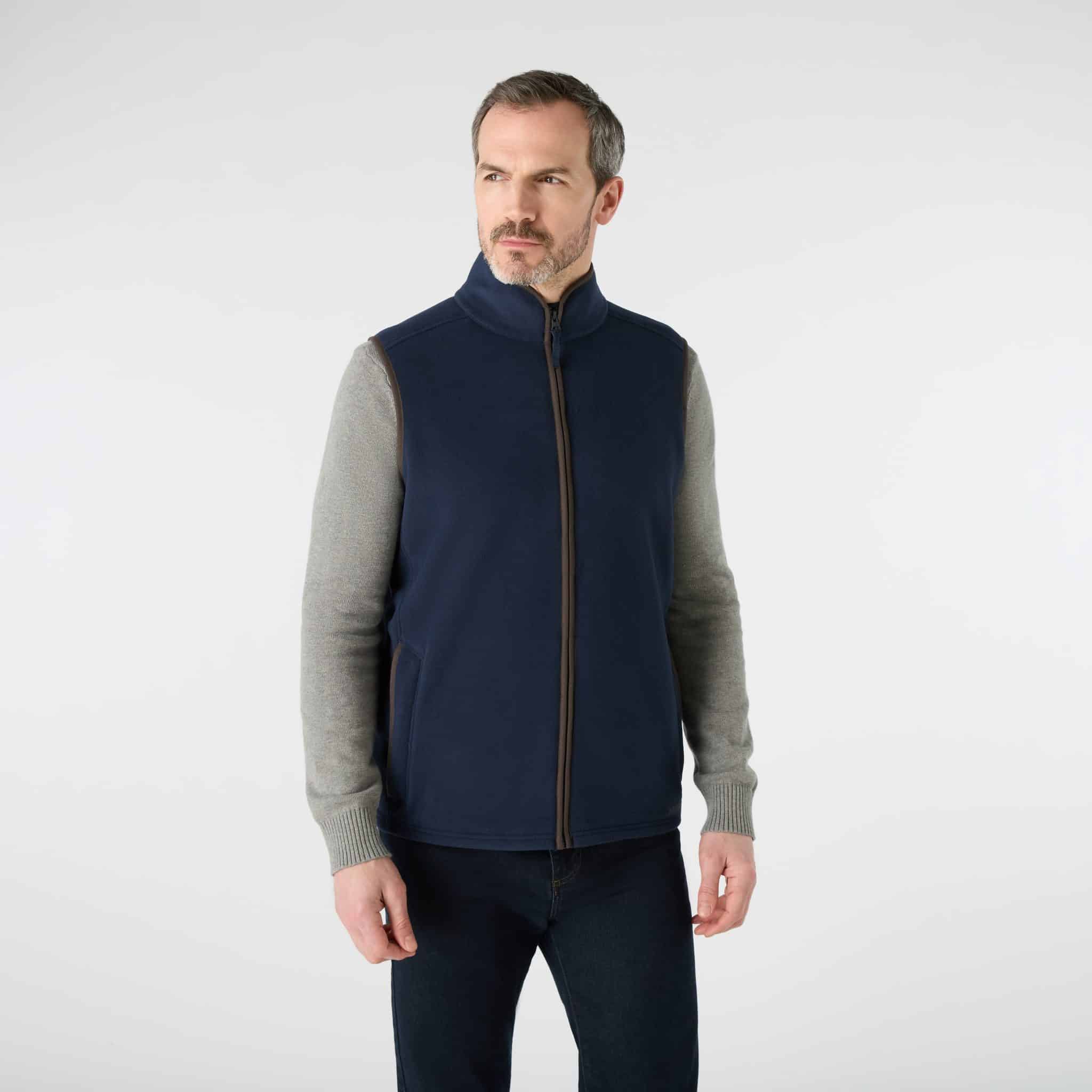 A man wearing a dark blue zip-up fleece gilet over a grey long-sleeved shirt and dark jeans.