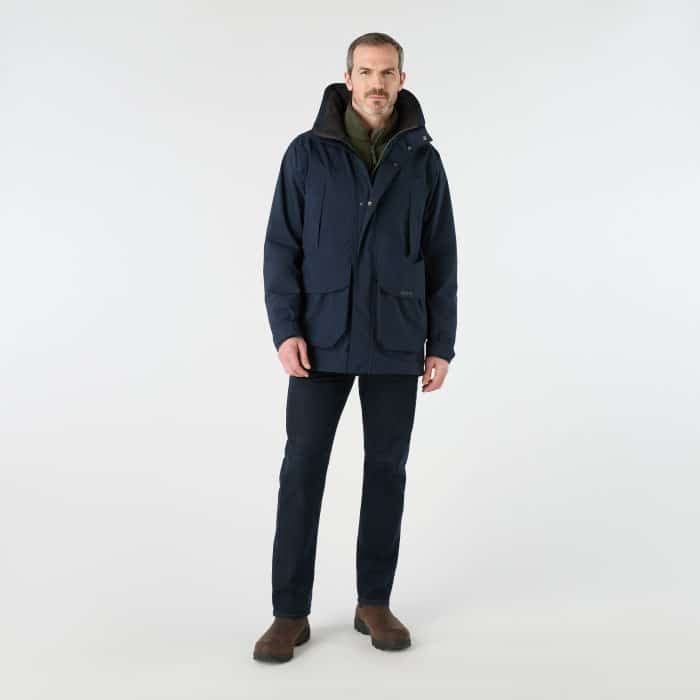 A man wearing a navy blue Musto waterproof jacket over an olive green fleece, dark jeans, and brown boots.