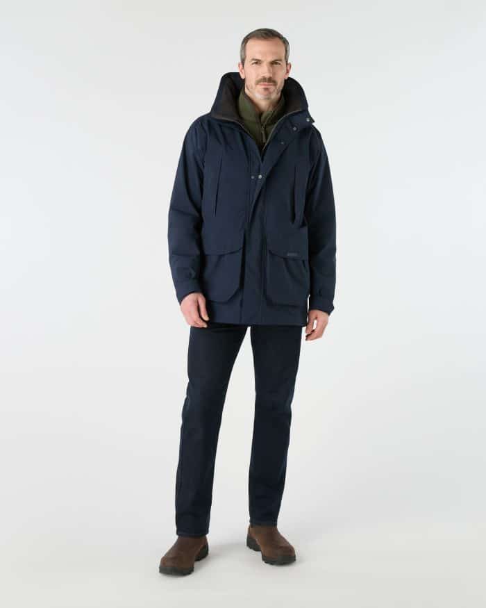 A man wearing a navy blue Musto waterproof jacket over an olive green fleece, dark jeans, and brown boots.