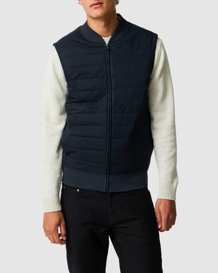 A man wearing a navy quilted zip-up gilet over a light-coloured long-sleeve jumper and black trousers.