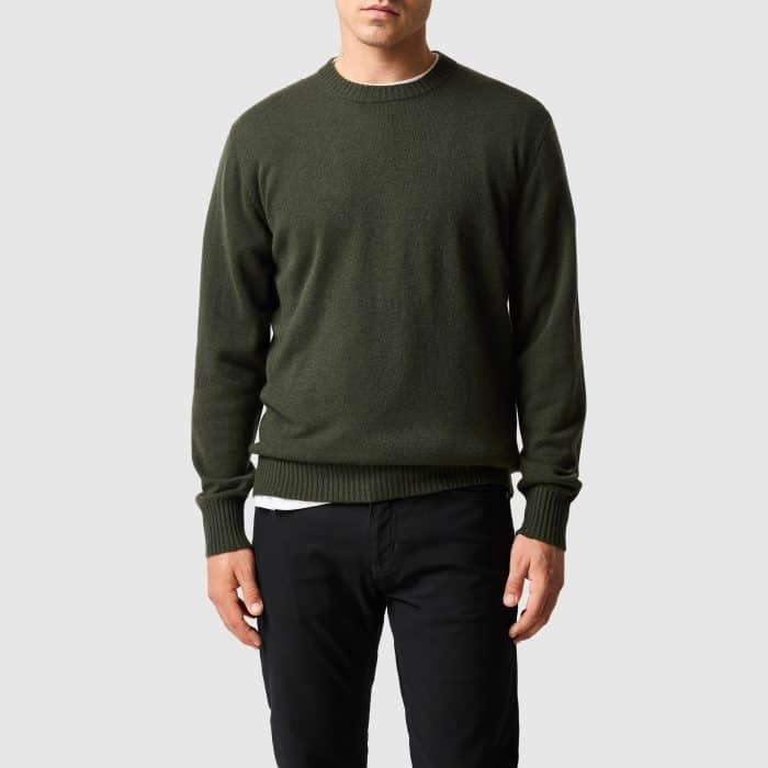 A person wearing a dark green crew neck jumper over a white T-shirt and black trousers.