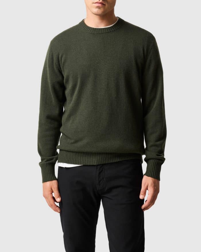 A person wearing a dark green crew neck jumper over a white T-shirt and black trousers.