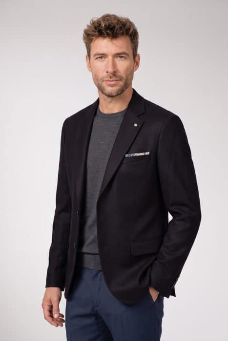 A man wearing a dark blazer over a grey jumper and blue trousers.