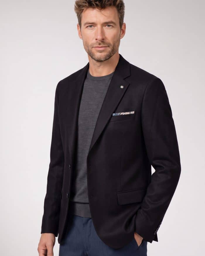 A man wearing a dark blazer over a grey jumper and blue trousers.