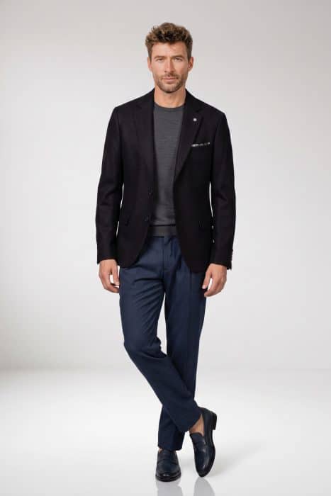 A man wearing a black blazer over a grey jumper, dark blue trousers, and navy loafers.