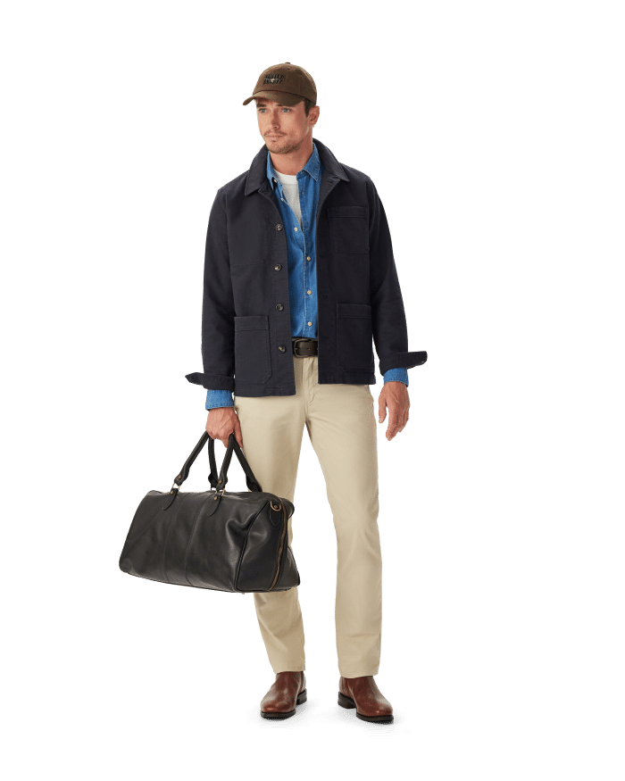 A man wearing a baseball cap, dark jacket, denim shirt, and khaki trousers carries a black leather holdall.