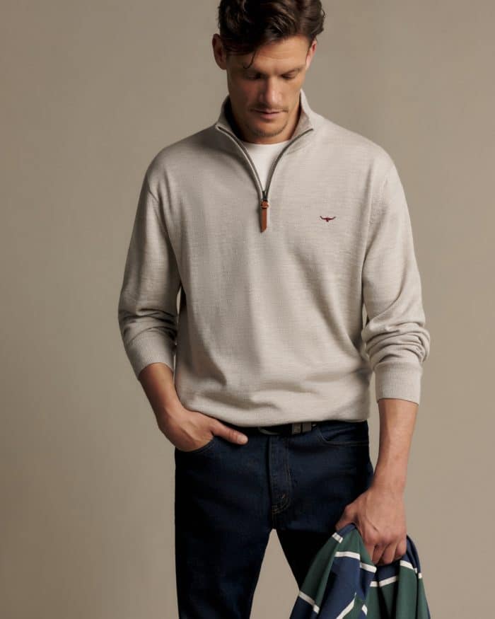A man wearing a beige quarter-zip jumper with a small red logo over a white T-shirt and dark blue jeans, holding a folded green, white, and navy striped shirt.