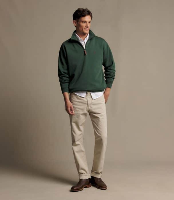 A man wears a dark green quarter-zip jumper over a white button-down shirt, paired with tan trousers and brown leather boots.