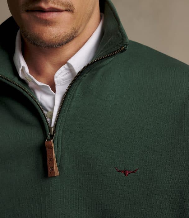 Close-up of a man wearing a dark green quarter-zip jumper with a brown leather zip pull and a small embroidered longhorn logo on the chest, layered over a white collared shirt.